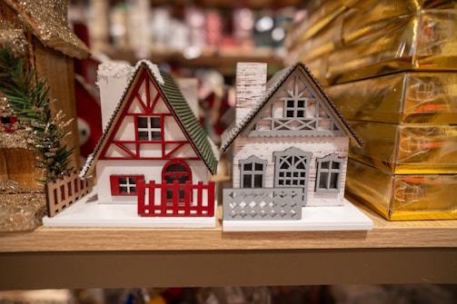 Two miniature wooden houses with a festive theme, featuring intricate details like fences and windows. The house on the left is predominantly white with red accents, including a red triangle pattern and matching fence. The house on the right is gray with a detailed cut-out design and a chimney, sitting beside golden gift boxes.