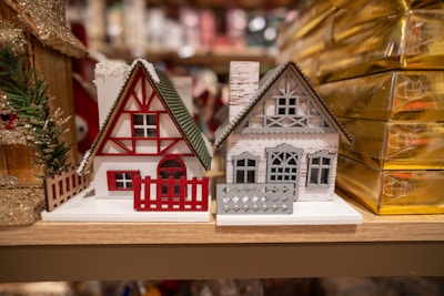 Two miniature wooden houses with a festive theme, featuring intricate details like fences and windows. The house on the left is predominantly white with red accents, including a red triangle pattern and matching fence. The house on the right is gray with a detailed cut-out design and a chimney, sitting beside golden gift boxes.