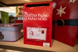 A red mailbox designed for letters to Santa Claus, 'Cartas Para O Papai Noel', is prominently displayed. It features a letter slot and an attached decorative envelope with a Santa illustration and address to the North Pole. The mailbox is part of a festive display with holiday-themed decorations, including tins and a snowflake design.