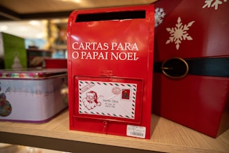 A red mailbox designed for letters to Santa Claus, 'Cartas Para O Papai Noel', is prominently displayed. It features a letter slot and an attached decorative envelope with a Santa illustration and address to the North Pole. The mailbox is part of a festive display with holiday-themed decorations, including tins and a snowflake design.