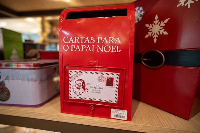 A red mailbox designed for letters to Santa Claus, 'Cartas Para O Papai Noel', is prominently displayed. It features a letter slot and an attached decorative envelope with a Santa illustration and address to the North Pole. The mailbox is part of a festive display with holiday-themed decorations, including tins and a snowflake design.