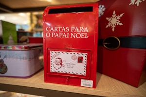 A red mailbox designed for letters to Santa Claus, 'Cartas Para O Papai Noel', is prominently displayed. It features a letter slot and an attached decorative envelope with a Santa illustration and address to the North Pole. The mailbox is part of a festive display with holiday-themed decorations, including tins and a snowflake design.
