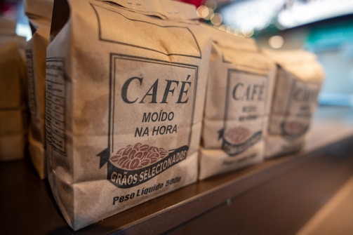 Several brown paper bags of coffee labeled with bold text 'Café Moído na Hora' and 'Grãos Selecionados' are neatly arranged on a shelf. The focus is on one bag, with others visible in soft focus in the background. The setting is likely a market or store with subtle lighting and a blurred backdrop.
