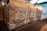 Several brown paper bags of coffee labeled with bold text 'Café Moído na Hora' and 'Grãos Selecionados' are neatly arranged on a shelf. The focus is on one bag, with others visible in soft focus in the background. The setting is likely a market or store with subtle lighting and a blurred backdrop.