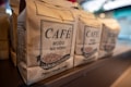 Several brown paper bags of coffee labeled with bold text 'Café Moído na Hora' and 'Grãos Selecionados' are neatly arranged on a shelf. The focus is on one bag, with others visible in soft focus in the background. The setting is likely a market or store with subtle lighting and a blurred backdrop.