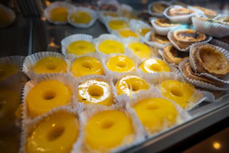 A variety of pastries displayed in a glass case, with a focus on vibrant yellow, custard-filled tarts neatly arranged in white paper cups. The tarts have a glossy surface and are accompanied by a stack of golden-brown pastries with flaky crusts in the background.