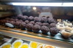 An assortment of chocolate truffles and other sweets displayed on a glass shelf. The chocolates are arranged in rows, with some topped with chocolate sprinkles. Nearby are white confections with chocolate centers. The background has a soft focus, highlighting the treats in the foreground.