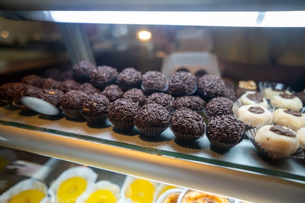 An assortment of chocolate truffles and other sweets displayed on a glass shelf. The chocolates are arranged in rows, with some topped with chocolate sprinkles. Nearby are white confections with chocolate centers. The background has a soft focus, highlighting the treats in the foreground.