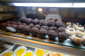 An assortment of chocolate truffles and other sweets displayed on a glass shelf. The chocolates are arranged in rows, with some topped with chocolate sprinkles. Nearby are white confections with chocolate centers. The background has a soft focus, highlighting the treats in the foreground.