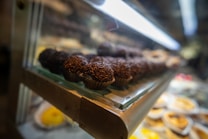 Several chocolate truffles with chocolate sprinkles are displayed in a glass case. They are placed in small paper cups, arranged in neat rows. The ambient light creates a warm and inviting atmosphere, highlighting the glossy texture of the sprinkles.