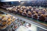 An assortment of desserts displayed in a glass case, featuring chocolate-covered treats topped with walnuts in the foreground and a variety of pastries like tarts and chocolate-covered strawberries in the background.