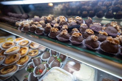 An assortment of desserts displayed in a glass case, featuring chocolate-covered treats topped with walnuts in the foreground and a variety of pastries like tarts and chocolate-covered strawberries in the background.