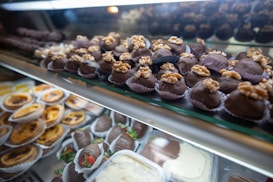 An assortment of desserts displayed in a glass case, featuring chocolate-covered treats topped with walnuts in the foreground and a variety of pastries like tarts and chocolate-covered strawberries in the background.