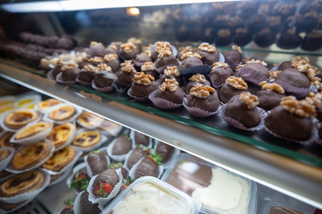 An assortment of desserts displayed in a glass case, featuring chocolate-covered treats topped with walnuts in the foreground and a variety of pastries like tarts and chocolate-covered strawberries in the background.