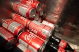 Several red cans with the Coca-Cola logo are stacked together inside a metallic container. Some bottles are also visible, suggesting a storage or cooling environment.