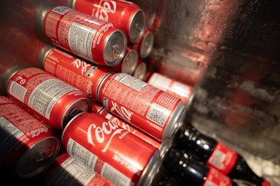 Several red cans with the Coca-Cola logo are stacked together inside a metallic container. Some bottles are also visible, suggesting a storage or cooling environment.