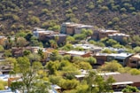 Several modern houses are nestled among a lush, green landscape with scattered trees and shrubbery. The homes are located at the base of a dry, rocky hill. The architecture features flat roofs and earth-toned exteriors, blending with the natural surroundings.