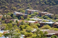 Several modern houses are nestled among a lush, green landscape with scattered trees and shrubbery. The homes are located at the base of a dry, rocky hill. The architecture features flat roofs and earth-toned exteriors, blending with the natural surroundings.