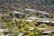 Several modern houses are nestled among a lush, green landscape with scattered trees and shrubbery. The homes are located at the base of a dry, rocky hill. The architecture features flat roofs and earth-toned exteriors, blending with the natural surroundings.