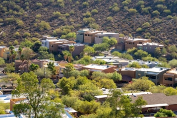 Several modern houses are nestled among a lush, green landscape with scattered trees and shrubbery. The homes are located at the base of a dry, rocky hill. The architecture features flat roofs and earth-toned exteriors, blending with the natural surroundings.
