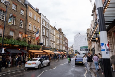 A bustling city street lined with historic buildings featuring a distinct European architectural style. Several people are walking along the sidewalk, some gathering near the entrance of a pub with a black and red awning. A French flag is prominently displayed on one of the buildings. Cars are parked along the street, and a person on a bicycle is riding in the bike lane. In the background, a large building under construction is covered in scaffolding and white sheeting. Overcast skies add to the urban atmosphere.