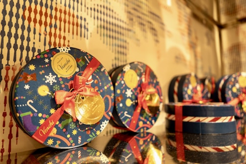 Several round, festive gift tins are displayed with a colorful design featuring holiday motifs like snowflakes, candy canes, and ornaments. Each tin is decorated with a bright red ribbon and a gold seal. The background consists of a textured pattern that complements the colors of the tins.