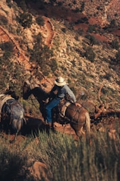 A person wearing a cowboy hat is riding a horse on a rugged trail in a desert landscape, with another horse laden with gear following closely. The trail is surrounded by rocks and sparse vegetation typical of arid environments.