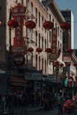 A busy street scene in what appears to be a Chinatown area, featuring numerous red lanterns hanging along the street, and signs in both Chinese and English. There are several shops, including a bakery and souvenir stores. People are walking along the sidewalk, and some are engaged in conversation. The architecture is characterized by brick buildings with fire escapes.