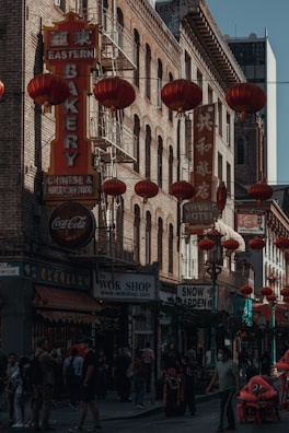 A busy street scene in what appears to be a Chinatown area, featuring numerous red lanterns hanging along the street, and signs in both Chinese and English. There are several shops, including a bakery and souvenir stores. People are walking along the sidewalk, and some are engaged in conversation. The architecture is characterized by brick buildings with fire escapes.