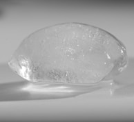 A clear, translucent ice cube with a smooth, rounded surface is placed on a white background. The light reflections and refractions create a textured effect within the ice.