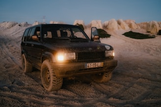 A dark, rugged off-road vehicle with its headlights on is parked on a sandy terrain with rocky formations in the background. The ground and vehicle are covered in a layer of grit, indicating recent off-road activity. One of the vehicle's doors is open.