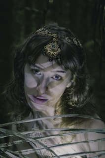 A woman with wavy hair adorned with an intricate, gem-encrusted headpiece stands amidst a shadowy, forest-like setting. Her expression appears calm yet intense, and beams of light create dramatic shadows across her face and the scene.