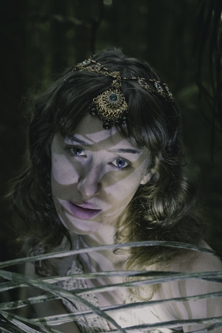 A woman with wavy hair adorned with an intricate, gem-encrusted headpiece stands amidst a shadowy, forest-like setting. Her expression appears calm yet intense, and beams of light create dramatic shadows across her face and the scene.