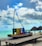 A small colorful boat with the word 'Preá' in bold, vibrant letters resting on top of it is placed on a wooden platform by the beach. The background features a serene ocean with a bright turquoise color under a partly cloudy sky. Two straw umbrellas are seen on the sand to the right, with a few people walking along the shoreline.