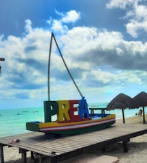 A small colorful boat with the word 'Pre&aacute;' in bold, vibrant letters resting on top of it is placed on a wooden platform by the beach. The background features a serene ocean with a bright turquoise color under a partly cloudy sky. Two straw umbrellas are seen on the sand to the right, with a few people walking along the shoreline.
