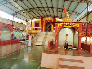 A serene temple interior with marble flooring and decorated pillars. At the center, a sacred altar displays a shivalinga with an ornate canopy adorned with marigold garlands. A devotee in traditional attire stands nearby, adding to the spiritual ambiance. Steps lead to a vibrant entrance built in traditional architecture with colorful railings.