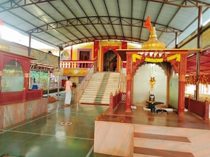 A serene temple interior with marble flooring and decorated pillars. At the center, a sacred altar displays a shivalinga with an ornate canopy adorned with marigold garlands. A devotee in traditional attire stands nearby, adding to the spiritual ambiance. Steps lead to a vibrant entrance built in traditional architecture with colorful railings.