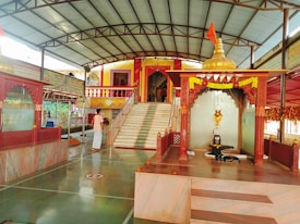 A serene temple interior with marble flooring and decorated pillars. At the center, a sacred altar displays a shivalinga with an ornate canopy adorned with marigold garlands. A devotee in traditional attire stands nearby, adding to the spiritual ambiance. Steps lead to a vibrant entrance built in traditional architecture with colorful railings.
