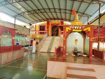A serene temple interior with marble flooring and decorated pillars. At the center, a sacred altar displays a shivalinga with an ornate canopy adorned with marigold garlands. A devotee in traditional attire stands nearby, adding to the spiritual ambiance. Steps lead to a vibrant entrance built in traditional architecture with colorful railings.