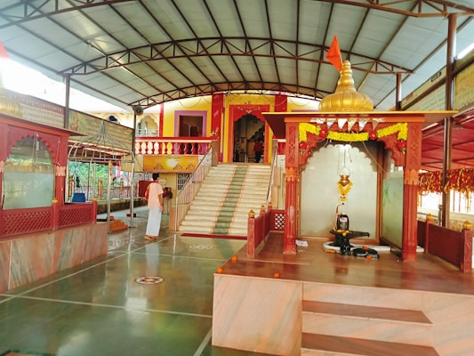 A serene temple interior with marble flooring and decorated pillars. At the center, a sacred altar displays a shivalinga with an ornate canopy adorned with marigold garlands. A devotee in traditional attire stands nearby, adding to the spiritual ambiance. Steps lead to a vibrant entrance built in traditional architecture with colorful railings.
