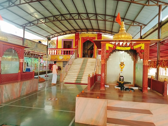 A serene temple interior with marble flooring and decorated pillars. At the center, a sacred altar displays a shivalinga with an ornate canopy adorned with marigold garlands. A devotee in traditional attire stands nearby, adding to the spiritual ambiance. Steps lead to a vibrant entrance built in traditional architecture with colorful railings.