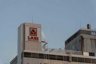 A tall, industrial-looking building with a sign reading 'LABI Hiroshima' is seen against a clear blue sky. The building has a modern design with metallic surfaces and several antennas on the roof.