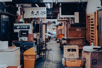 A narrow alleyway lined with various signs in Japanese characters. There are stacks of boxes and crates on the sides, alongside shelves and other objects. The area appears cluttered yet organized with a hint of bustling activity further down the alley.