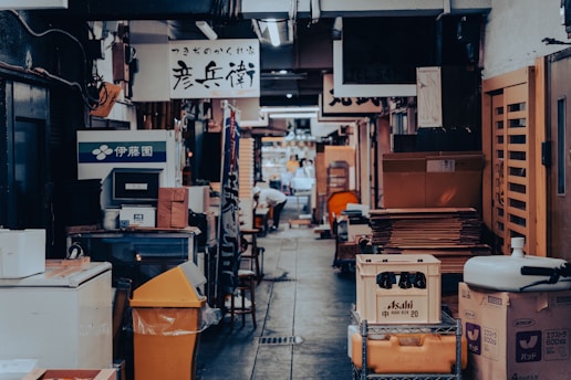 A narrow alleyway lined with various signs in Japanese characters. There are stacks of boxes and crates on the sides, alongside shelves and other objects. The area appears cluttered yet organized with a hint of bustling activity further down the alley.