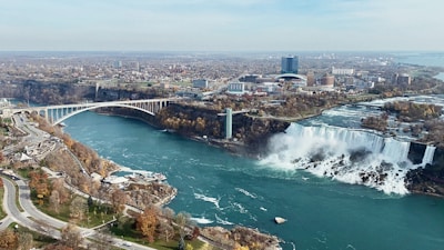 An aerial view of Niagara Falls with a waterfall prominently featured on the right side of the image. A bridge extends over the river, connecting two areas of land. To the left, there are roads, pathways, and green spaces with trees that have autumnal foliage. In the background, there is an urban landscape with buildings and infrastructure.