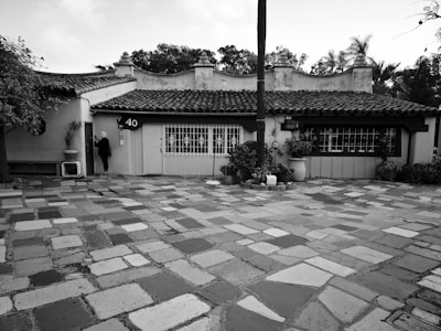 A single-story house with a tiled roof and colonial architectural style, surrounded by lush vegetation. The patio features a rustic, uneven stone pavement, and there is a large, tall tree in front of the house. A person is standing at the entrance, which has the number 40 displayed.
