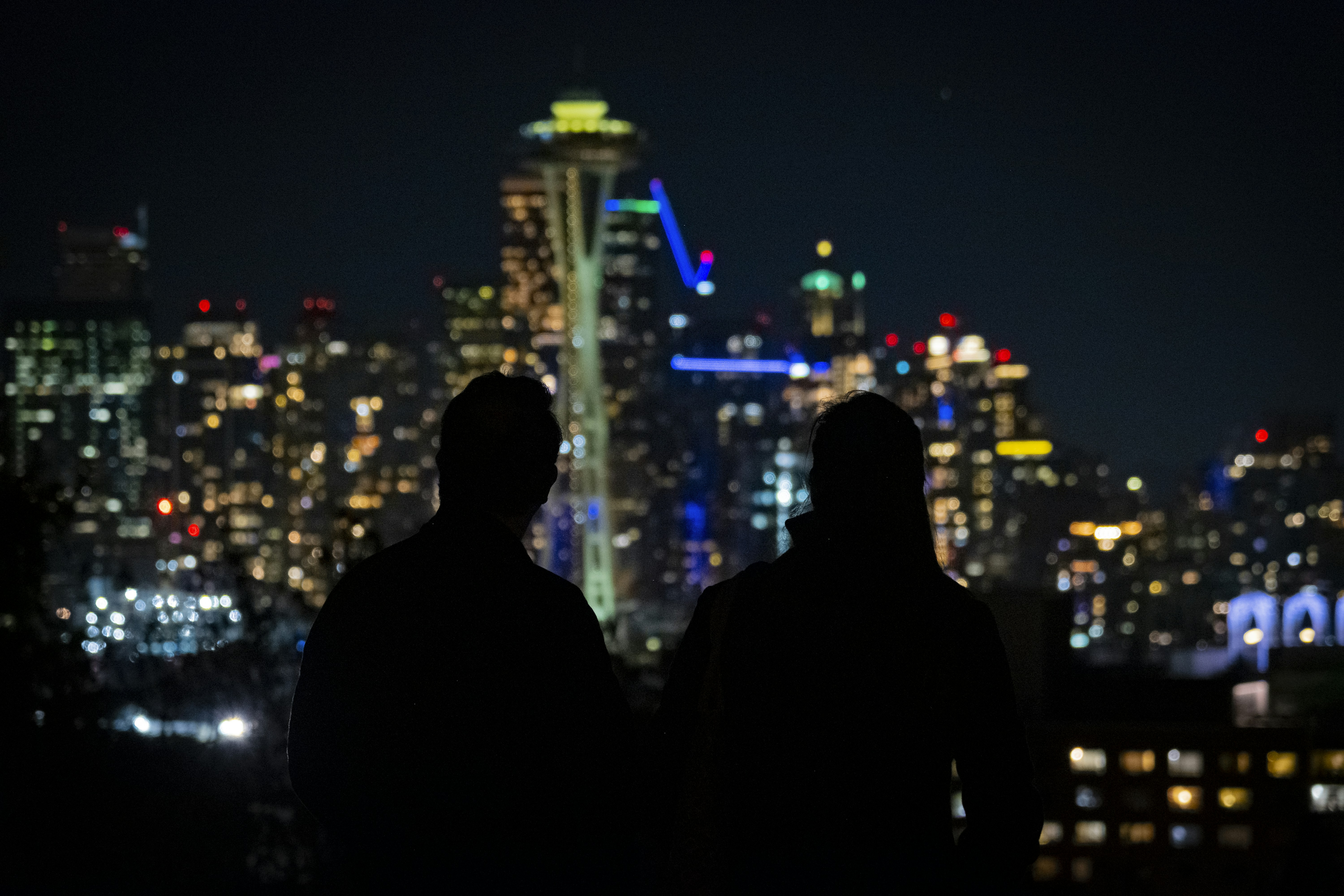a couple of men standing next to each other in front of a city, A silhouette of a couple looking at Seattle Skyline on a cold November evening
