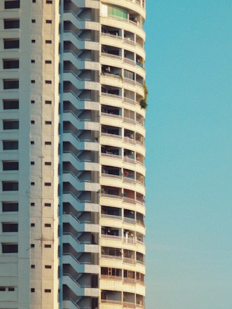 A tall, modern apartment building with numerous balconies and a clean, minimalist design. The structure features prominent vertical lines enhanced by shadows, with a clear blue sky in the background. One balcony has green vegetation visible.