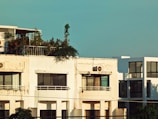 A modern, multi-story apartment building with a rooftop garden filled with greenery. The facade is slightly weathered with visible air conditioning units. Large windows allow natural light into the rooms. The sky in the background is clear and blue.