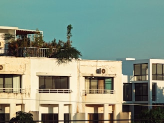 A modern, multi-story apartment building with a rooftop garden filled with greenery. The facade is slightly weathered with visible air conditioning units. Large windows allow natural light into the rooms. The sky in the background is clear and blue.